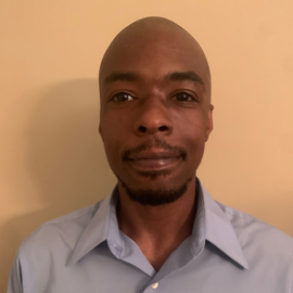 A candid self taken photo of Logan Willoughby wearing a light blue collared shirt against a tan wall.